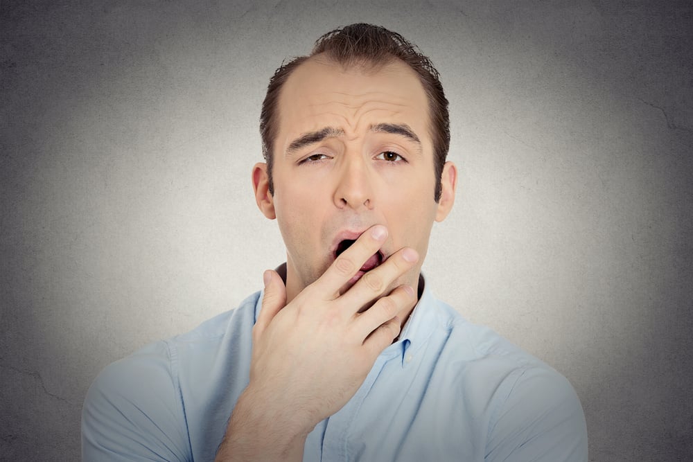 Closeup portrait headshot sleepy young businessman funny guy placing hand on mouth yawning looking at camera isolated grey wall background. Negative human emotion face expression feeling body language